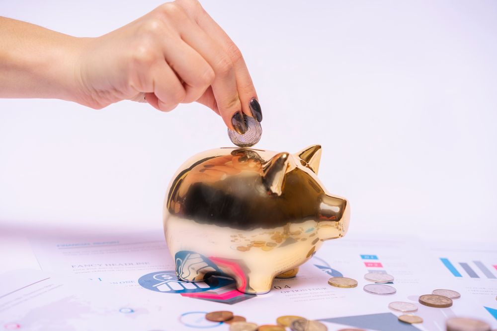 Hand wirft eine Münze in ein goldenes Sparschwein neben Finanzunterlagen